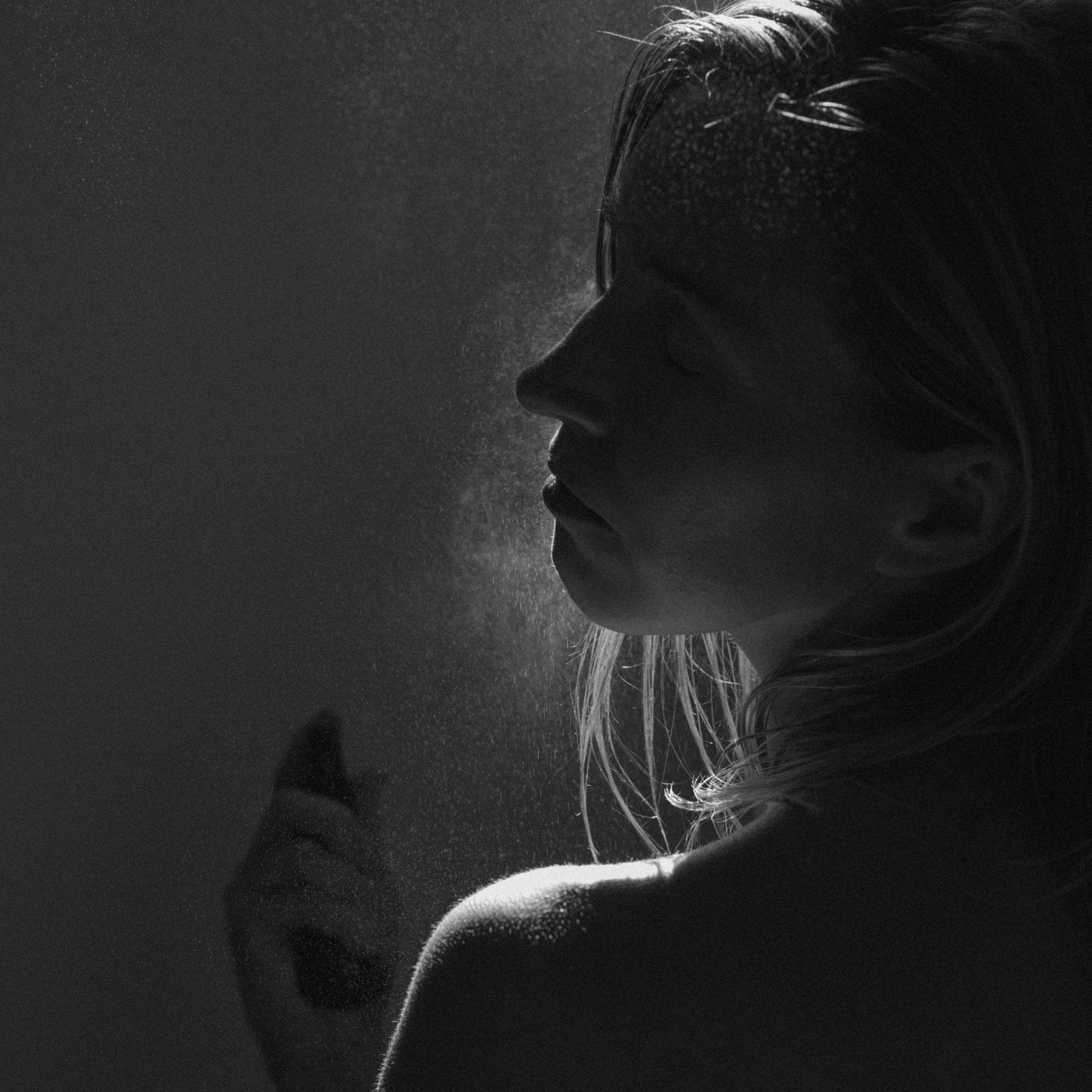 A woman in profile sprays Summer Solace Tallow's Jasmine Sambac + Magnolia Perfume Mist – Water-Based onto her face, with soft lighting highlighting the mist’s droplets and casting dramatic shadows across her features.