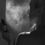 A person sprays Summer Solace Tallow’s Jasmine Sambac + Magnolia Perfume Mist – Water-Based onto their face in black and white, dramatic lighting highlighting the floral mist and their profile.