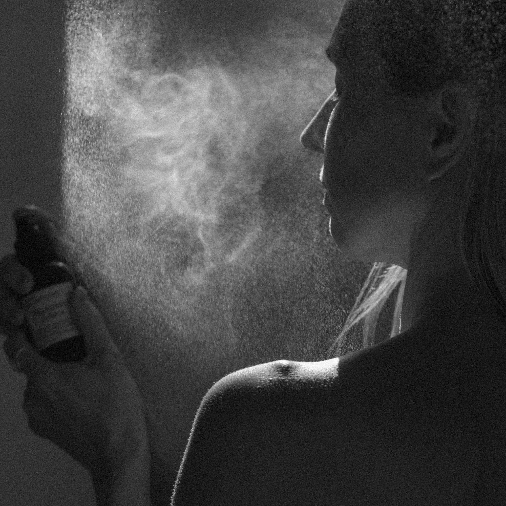 A person sprays Summer Solace Tallow’s Jasmine Sambac + Magnolia Perfume Mist – Water-Based onto their face in black and white, dramatic lighting highlighting the floral mist and their profile.