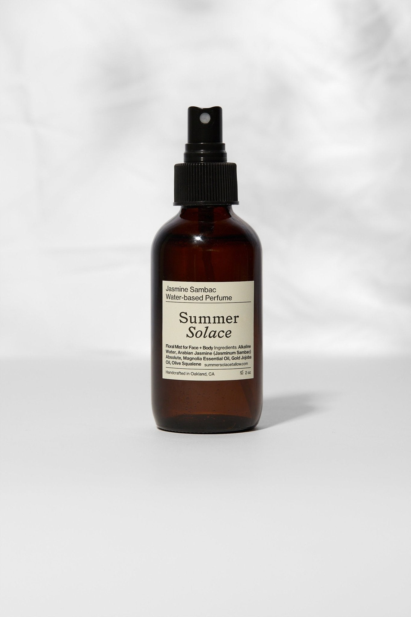 A brown glass spray bottle labeled "Jasmine Sambac + Magnolia Perfume Mist – Water-Based" by Summer Solace Tallow stands upright on a light surface with a white, softly textured background, highlighting this delicate floral perfume.