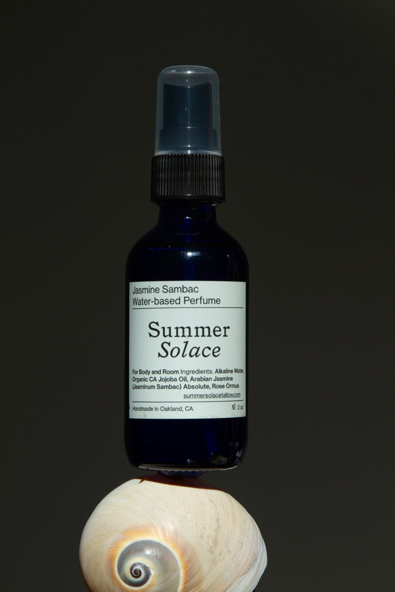 A dark blue spray bottle labeled "Summer Solace Tallow Jasmine Sambac + Magnolia Perfume Mist – Water-Based"—a delicate floral fragrance—rests atop a white seashell against a plain dark background.