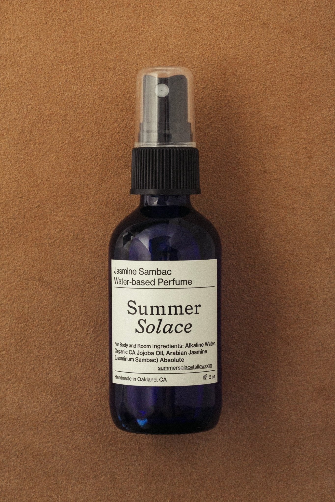 A dark blue spray bottle labeled "Summer Solace Tallow" Jasmine Sambac + Magnolia Perfume Mist sits on a tan surface. This water-based perfume is handcrafted in Oakland, CA, with ingredients listed on the label.