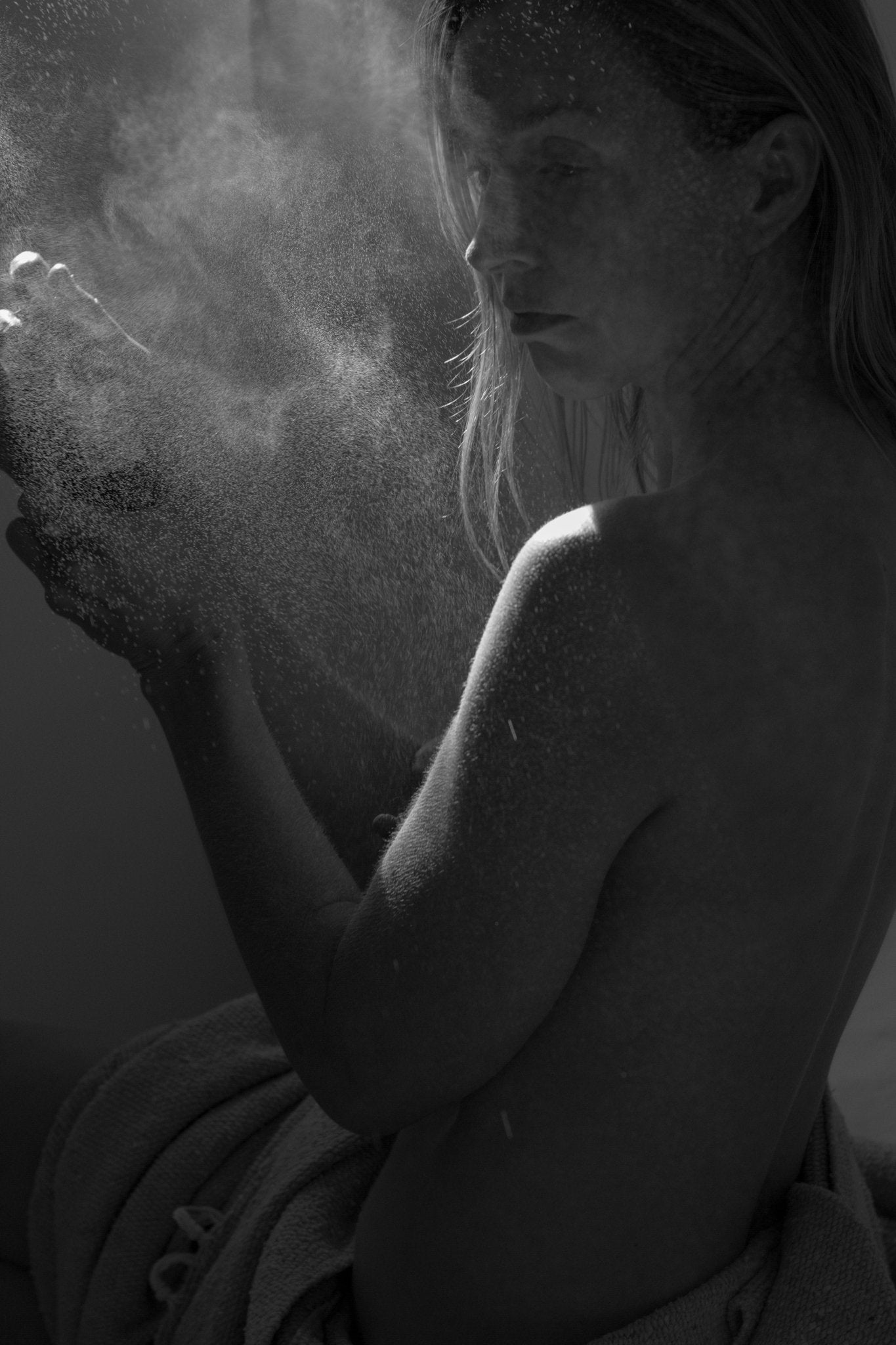 Bathed in soft light, a woman draped in a towel spritzes Summer Solace Tallow's Jasmine Sambac + Magnolia Perfume Mist as delicate droplets swirl around her, creating an ethereal and dramatic atmosphere.