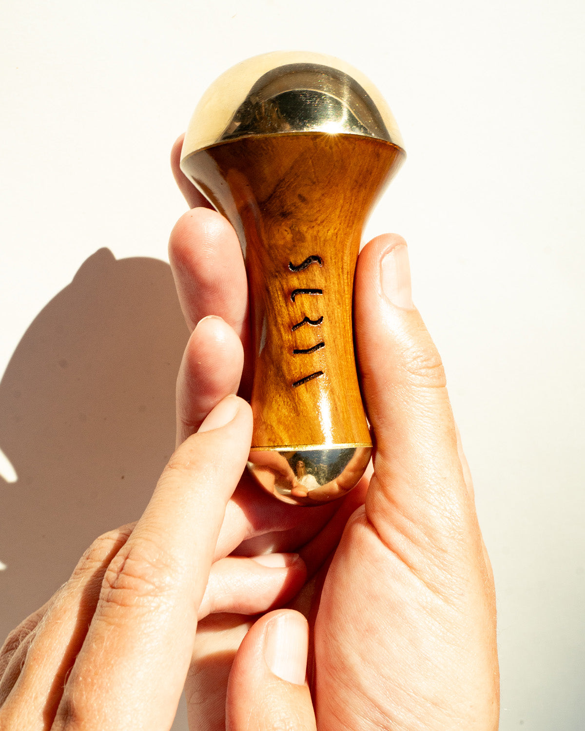 A person holds a Surya Kansa Wand, a wooden and gold-toned massage tool made from skin-healing metal with "SEED" engraved vertically on its side, set against a plain light background.