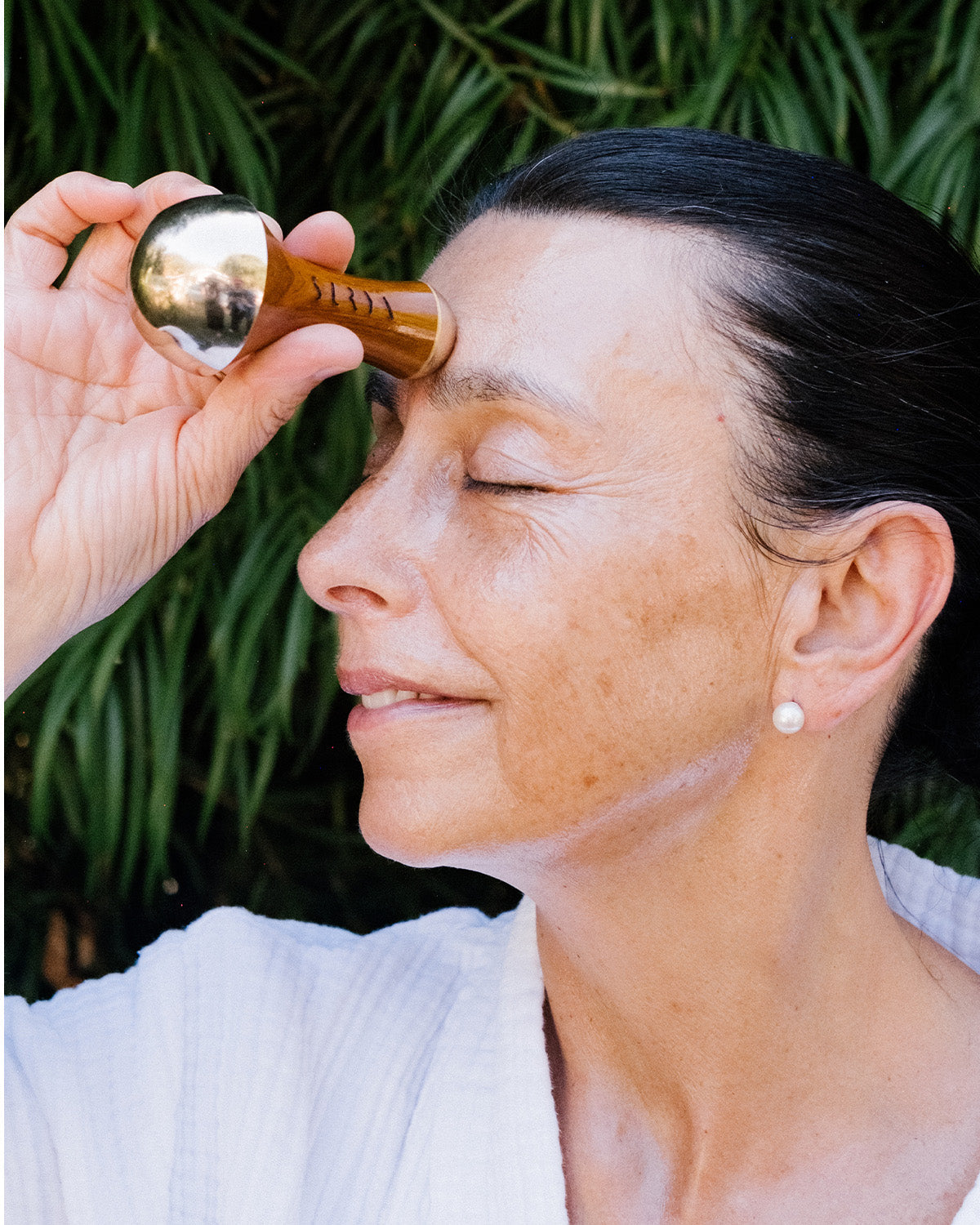 A smiling woman in a white robe, eyes closed, uses the Surya Kansa Wand—an Ayurvedic skincare tool made from healing metal—on her forehead, standing before lush green plants.