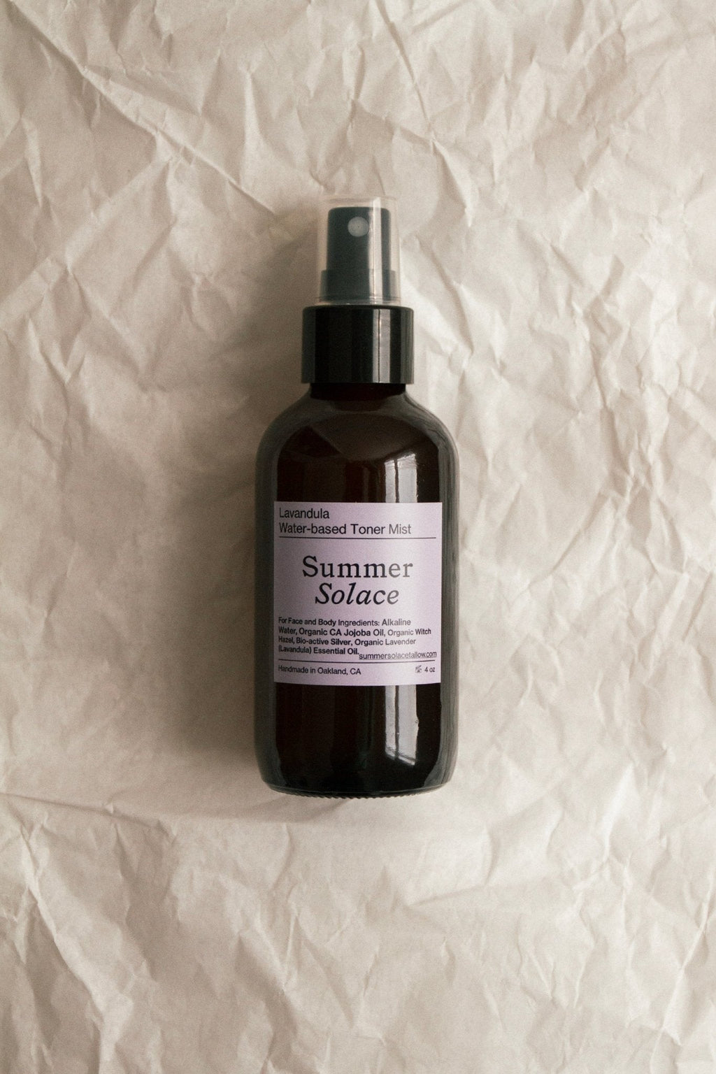 A dark spray bottle labeled "Summer Solace Tallow Lavender & White Lotus Toner – Organic Floral Mist" rests on crumpled, off-white paper. The label includes product details and an Oakland, CA address.
