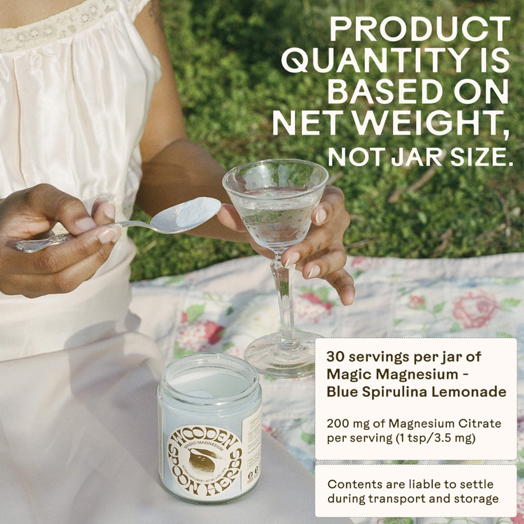 A person sits on a picnic blanket holding a glass beside an open jar labeled "WOODEN SPOON HERBS Magic Magnesium - Blue Spirulina Lemonade." Text notes product quantity is by net weight, not jar size. Serving and ingredient info provided—Magnesium Citrate supports calm and energy.