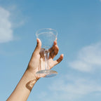 A hand holds up a Magic Magnesium bottle from WOODEN SPOON HERBS against a calm, clear blue sky with wispy clouds, evoking energy and tranquility.