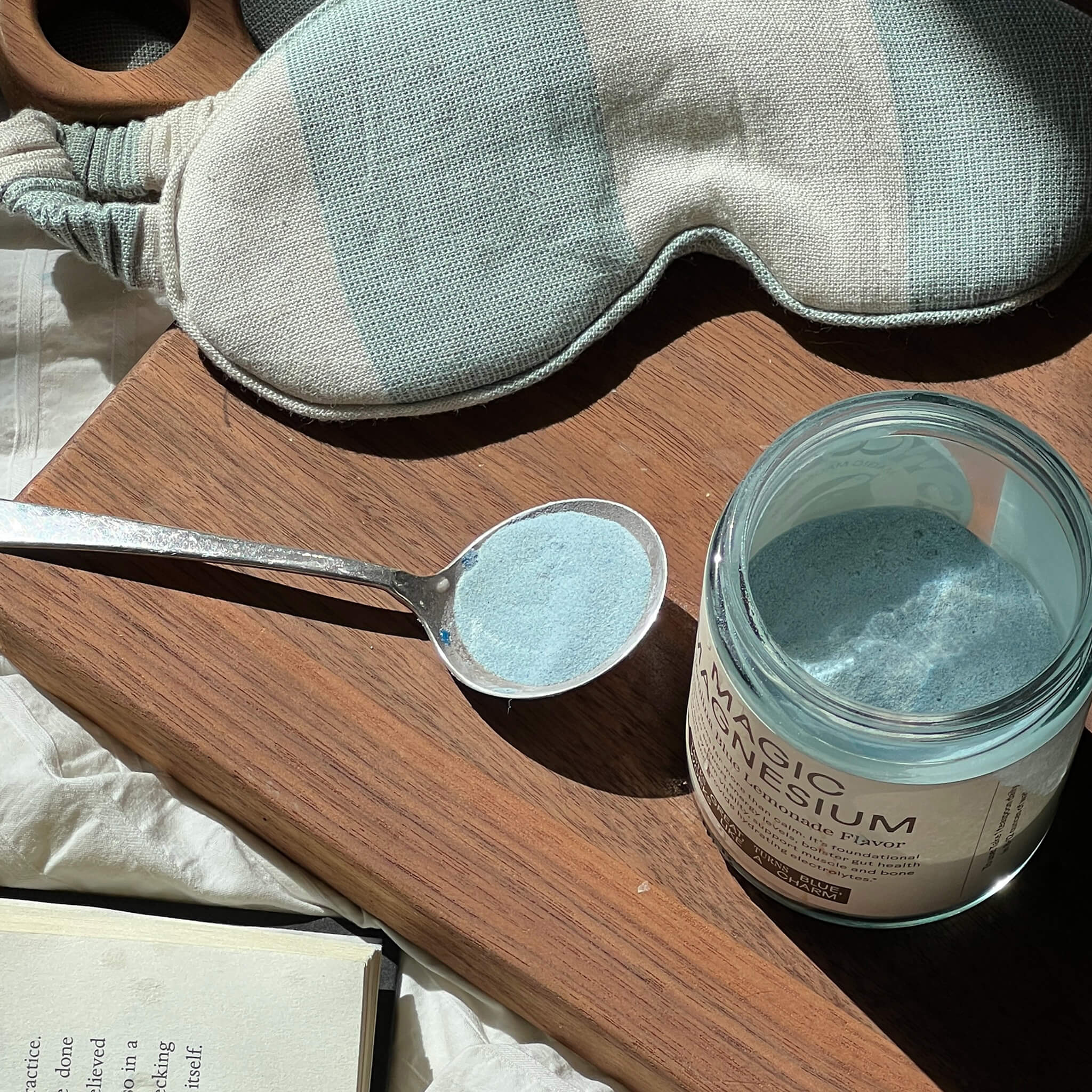 A spoon with blue powder sits next to an open WOODEN SPOON HERBS Magic Magnesium jar on a wooden tray. Sunlight highlights a striped sleep mask and book, enhancing the calm mood inspired by this magnesium supplement.