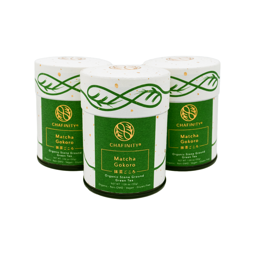 Three cylindrical containers of Chafinity Tea's Organic Stone Ground Ceremonial Matcha Gokoro are displayed against a black background. The containers feature a green and white design with gold detailing, embodying the essence of Japanese tea ceremonies. They are labeled as organic, stone-ground matcha green tea powder.
