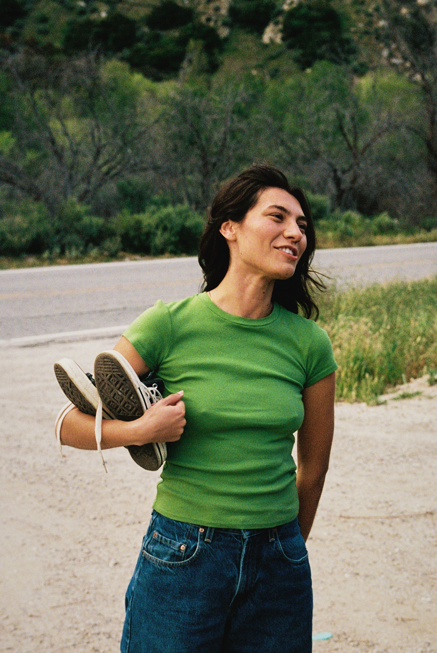 A woman in a green LA Relaxed Bebe Organic Tee and jeans smiles by the roadside, holding sneakers. Lush greenery and a winding road extend behind her.