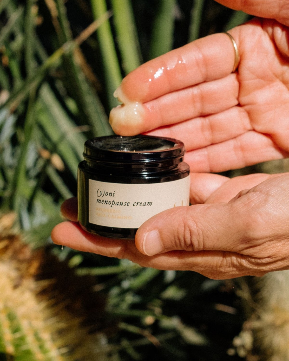 A hand holds an open jar of Surya's (y)oni Menopause Cream infused with Ayurvedic botanicals, while another hand scoops a small amount with a fingertip. The background features blurred greenery, highlighting the intimate skincare routine.