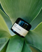 A black jar labeled "Surya (y)oni Menopause Cream" rests on succulent green leaves—an intimate skin moisturizer inspired by Ayurvedic botanicals.