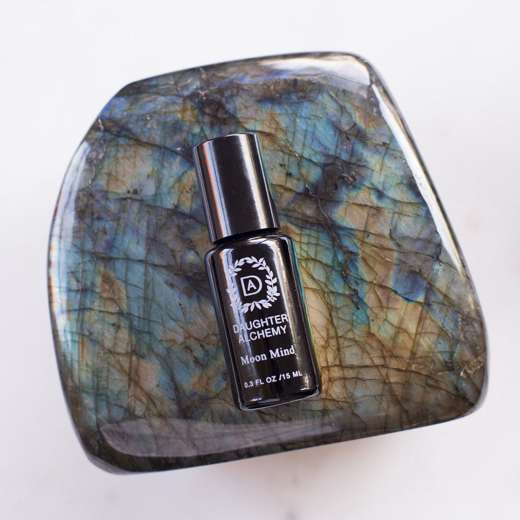 A small black bottle of Daughter Alchemy Moon Mind® Botanical Intuition Oil rests on a polished iridescent stone, its exquisite meditative blend expertly infused with organic jasmine and ylang ylang oils.