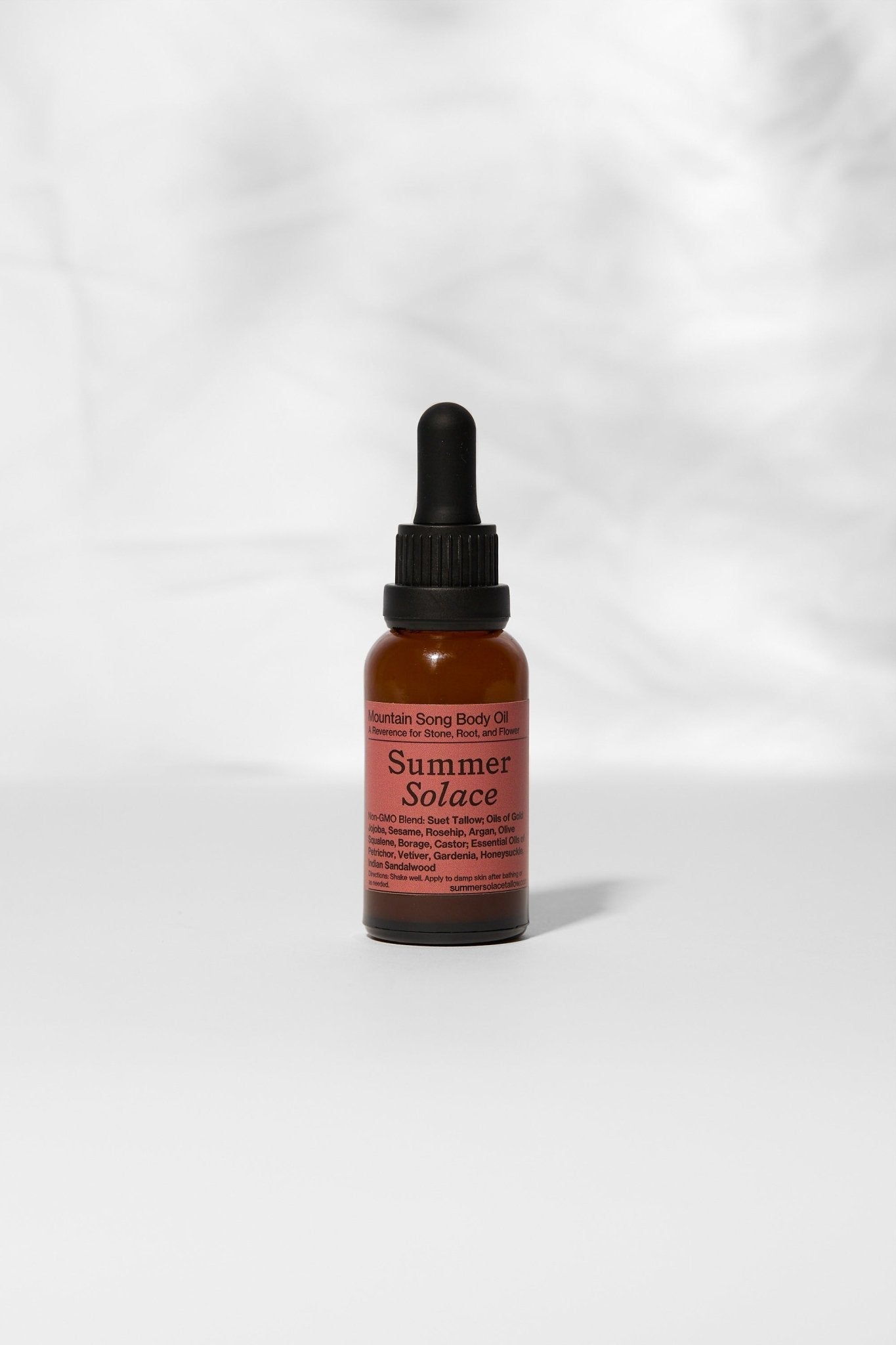 A small brown glass dropper bottle with a black cap and a pink "Summer Solace Tallow" label stands upright against a white background, featuring Mountain Song Body and Hair Oil – Petrichor, Gardenia, Honeysuckle (Limited Edition).