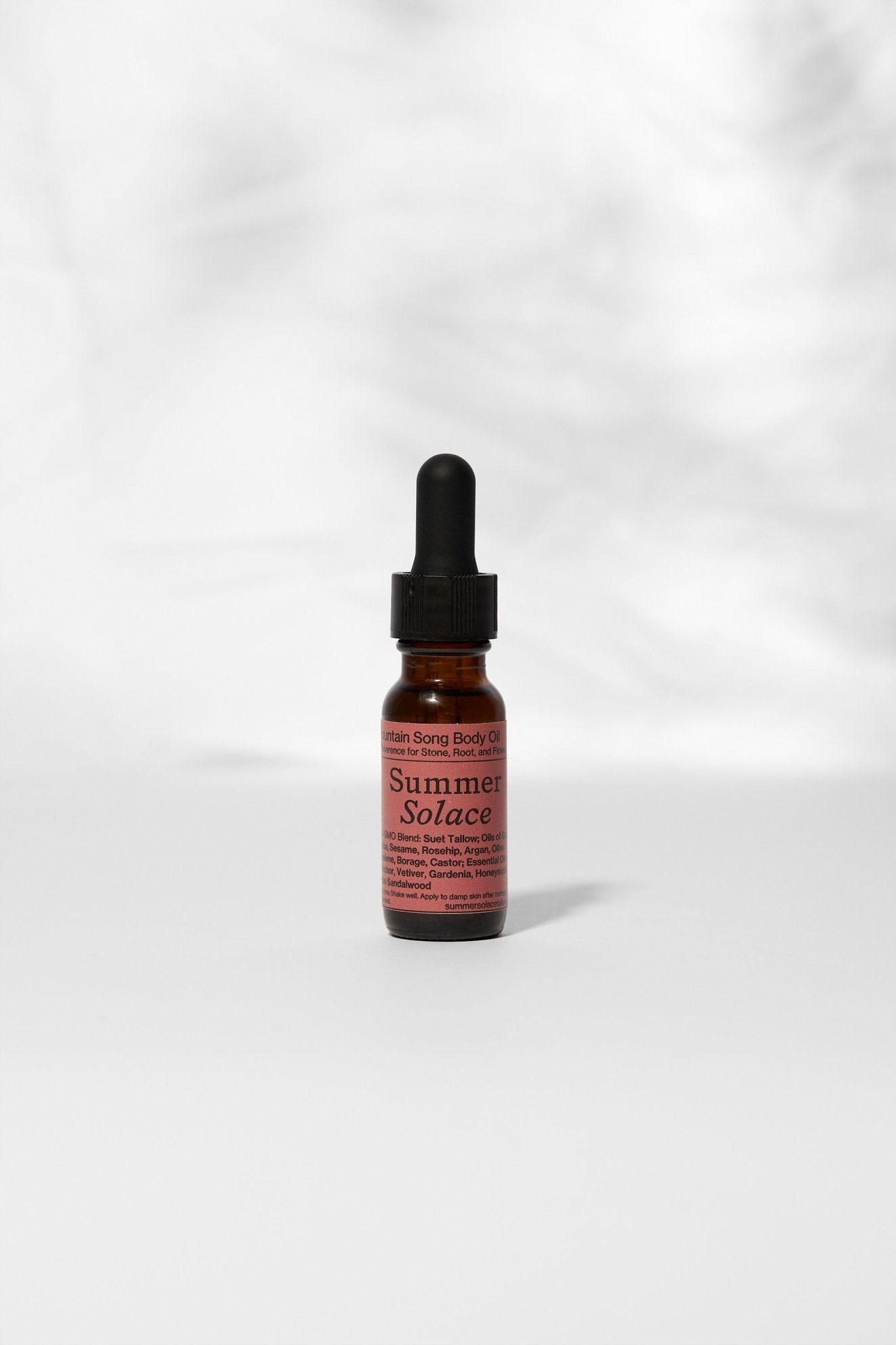 A small amber glass dropper bottle labeled "Summer Solace Tallow" stands upright on a light surface, displaying the limited edition Mountain Song Body and Hair Oil in Petrichor, Gardenia, Honeysuckle with soft shadows behind.