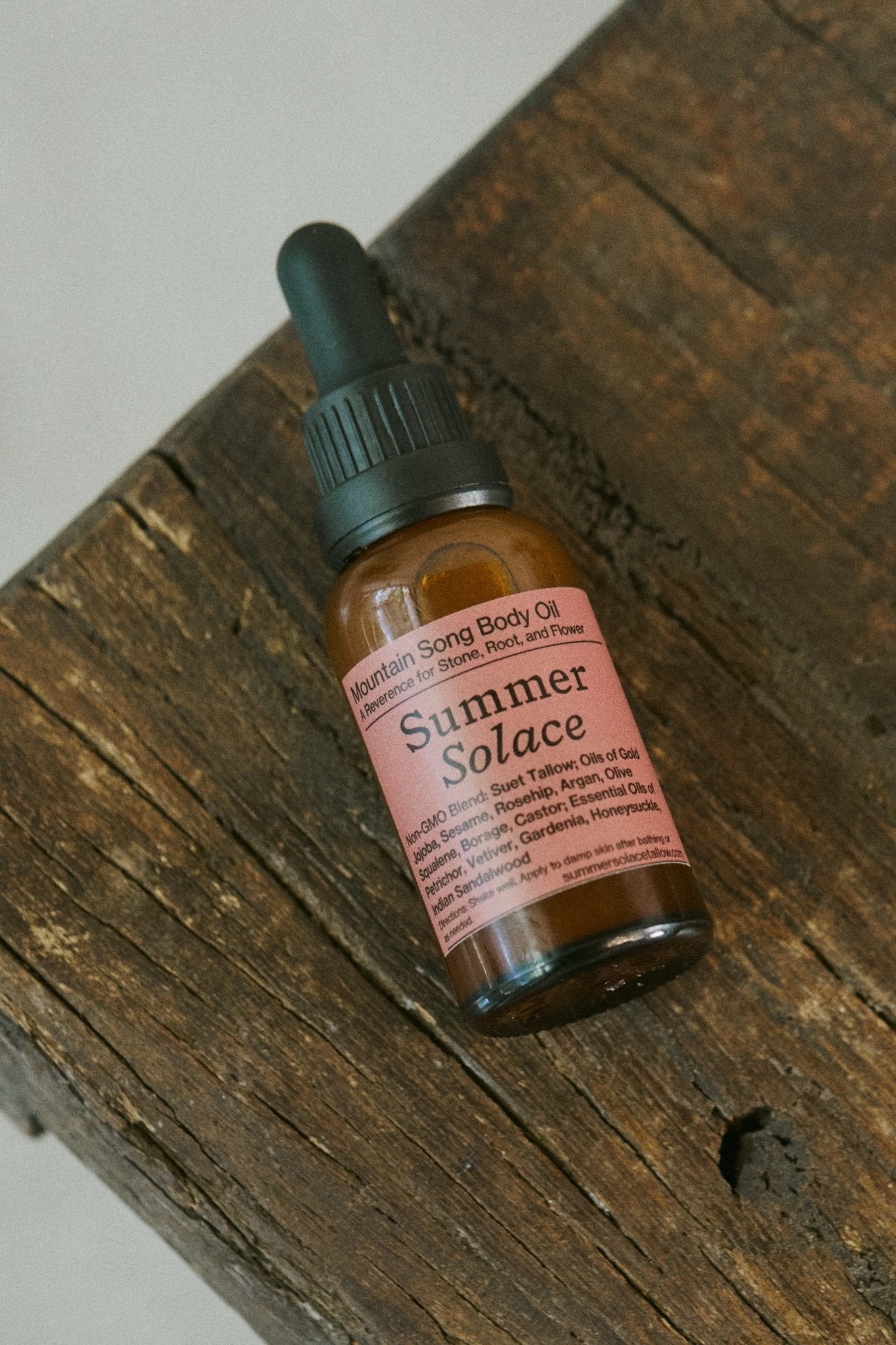 A brown glass dropper bottle labeled "Mountain Song Body and Hair Oil – Petrichor, Gardenia, Honeysuckle (Limited Edition)" by Summer Solace Tallow sits on a rustic wooden surface, its pink label softly glowing.