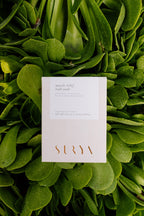 A white box labeled "Muscle Relief Bath Soak" by Surya sits on lush green leaves. Its minimalist design features gold "Surya" text, capturing the restorative essence of a spa therapy experience.