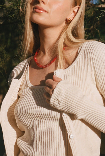 A woman in a cream LA RELAXED Pointelle Cardigan holds her sweater. She wears a ribbed knit top with pearlescent buttons, straight blonde hair, a red beaded necklace, and gold hoops as sunlight highlights her face and neck.