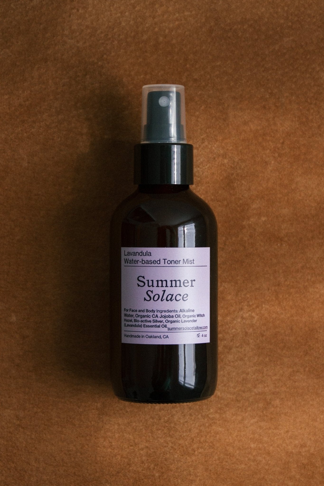 A dark amber spray bottle labeled "Lavender & White Lotus Toner – Organic Floral Mist" by Summer Solace Tallow rests on a brown suede surface, with a label noting it is handcrafted in Oakland, CA and contains organic ingredients.