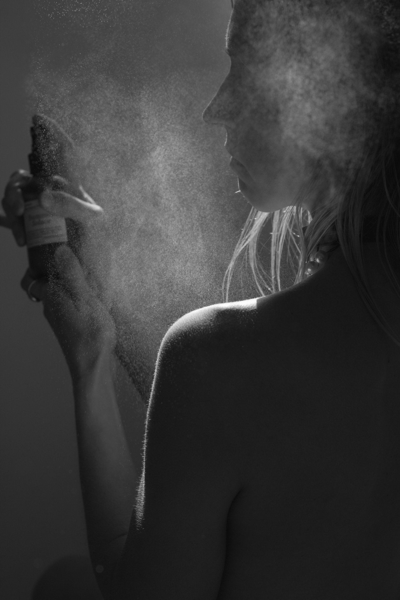 A woman sprays Summer Solace Tallow's Lavender & White Lotus Toner – Organic Floral Mist onto her face and shoulder, visible fine mist particles catching the soft light and highlighting her profile and bare skin.