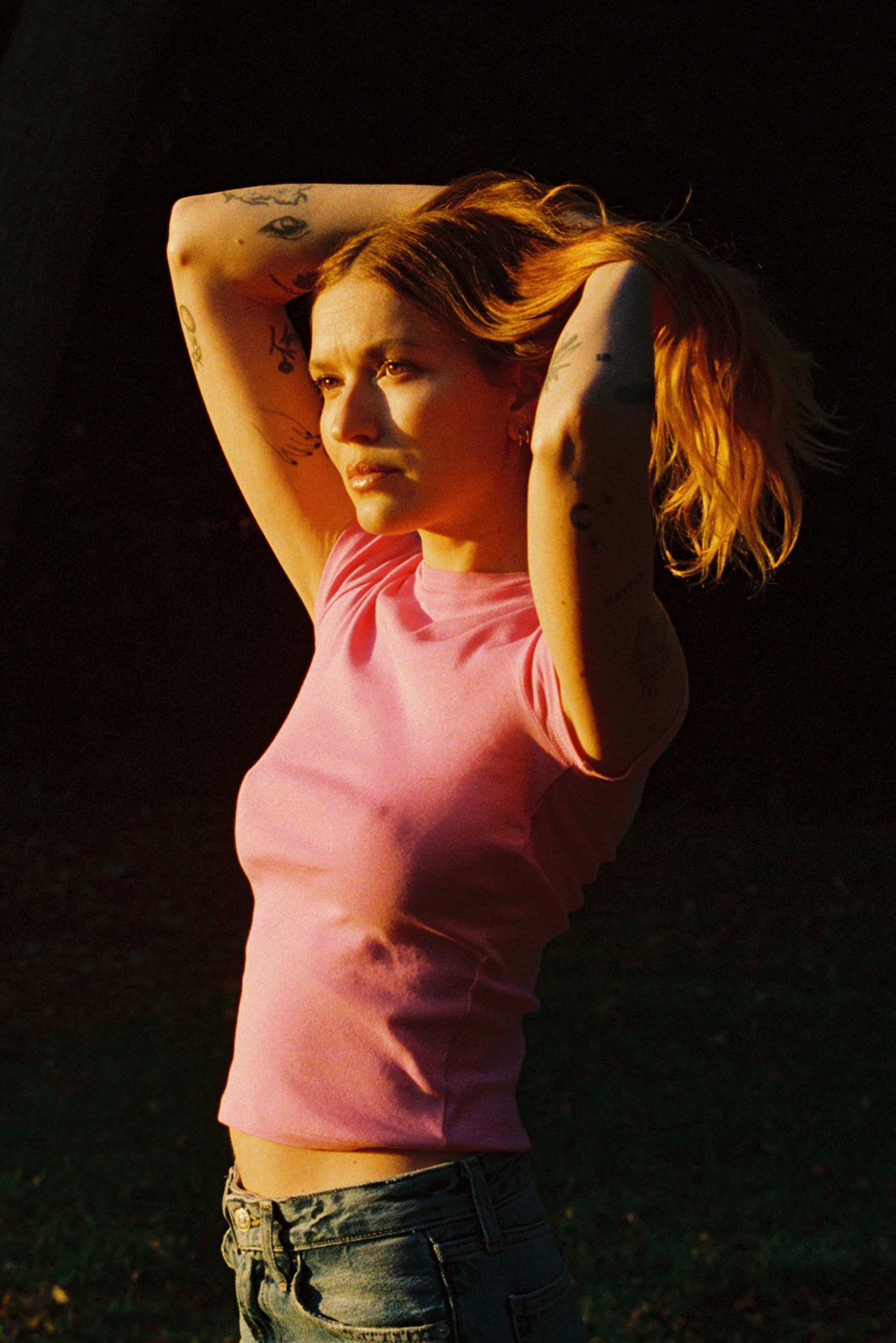 A tattooed person with long hair in a bun stands outside in warm sunlight, wearing a pink Bebe Organic Tee by LA Relaxed and jeans, gazing thoughtfully into the distance against a dark grassy background.