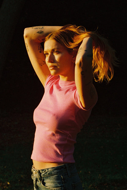 A tattooed person with long hair in a bun stands outside in warm sunlight, wearing a pink Bebe Organic Tee by LA Relaxed and jeans, gazing thoughtfully into the distance against a dark grassy background.