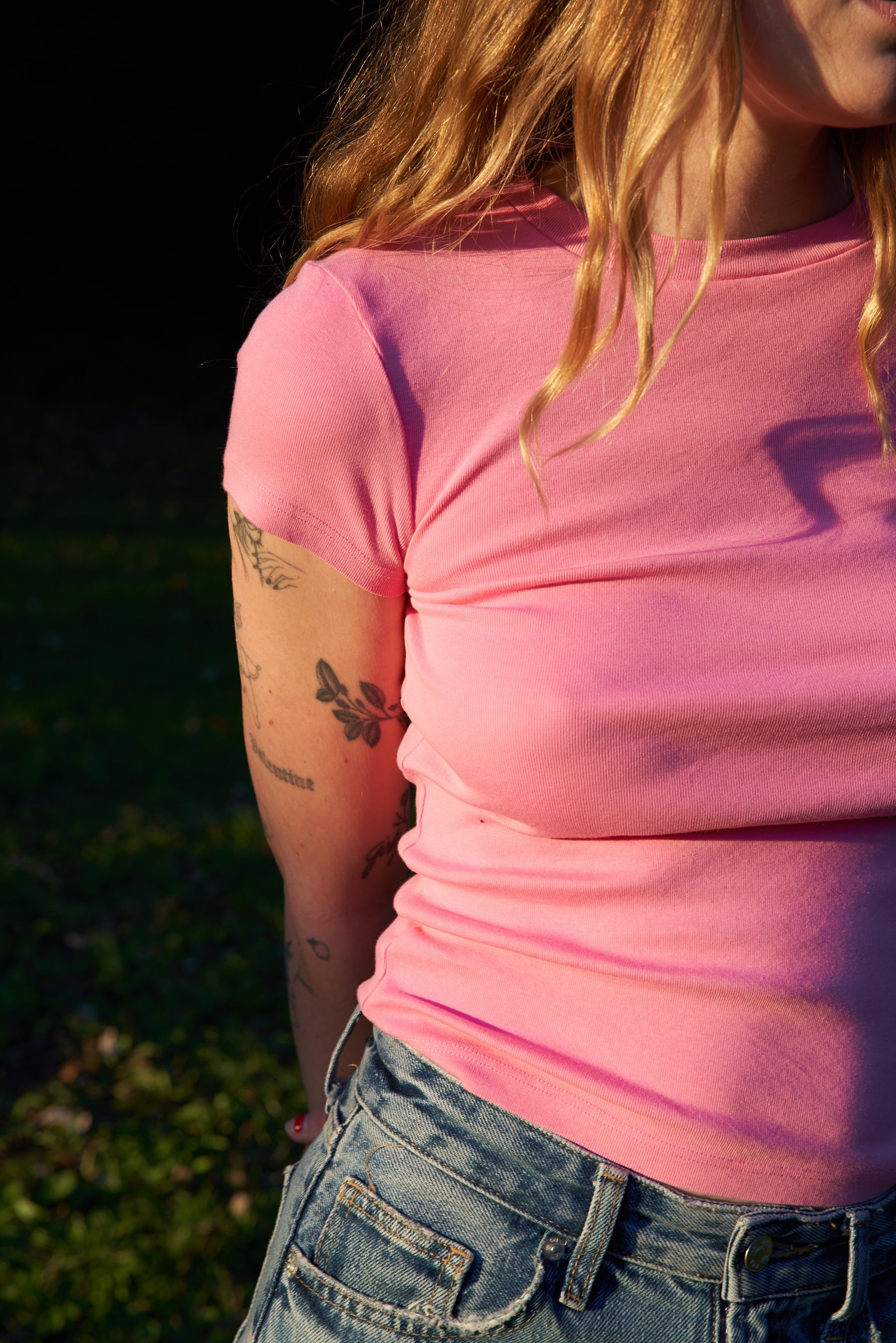 A person with long wavy hair is outdoors, wearing a pink LA Relaxed Bebe Organic Tee and blue denim shorts. Their tattooed arms are partially in shadow, while sunlight highlights the organic cotton fabric of their clothing.