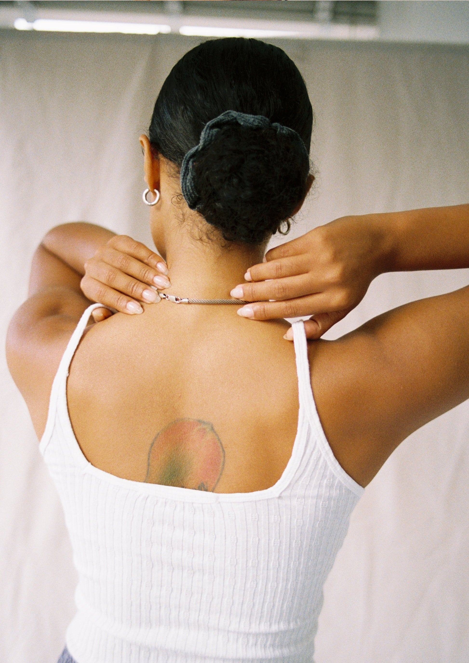 A person with a peach tattoo on their upper back adjusts a necklace, wearing the LA Relaxed Pointelle Square Cami. Their dark hair is pulled back with a scrunchie. The photo is taken from behind against a light background.