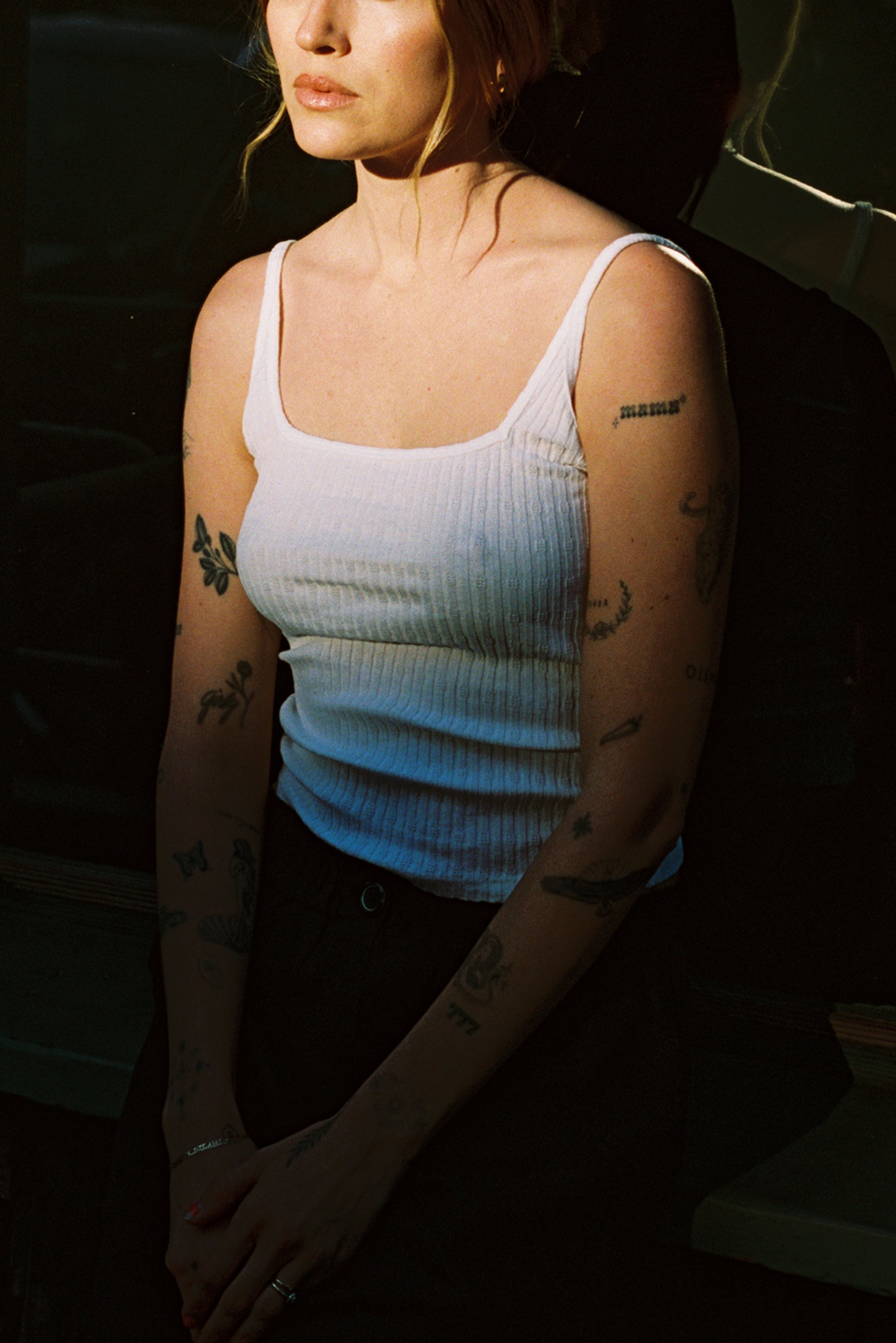 A woman with tattoos on her arms wears the LA Relaxed Pointelle Square Cami and dark pants, standing partially in shadow with her hands clasped. Her face is partly out of the frame.