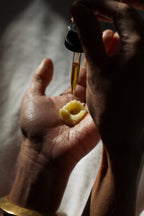 A hand holds a dollop of cream as Emanations Profound Face Oil by Summer Solace Tallow—featuring tallow, emu oil, champa, tuberose & white ginger lily—is dispensed from a dropper above, sunlight casting gentle shadows.