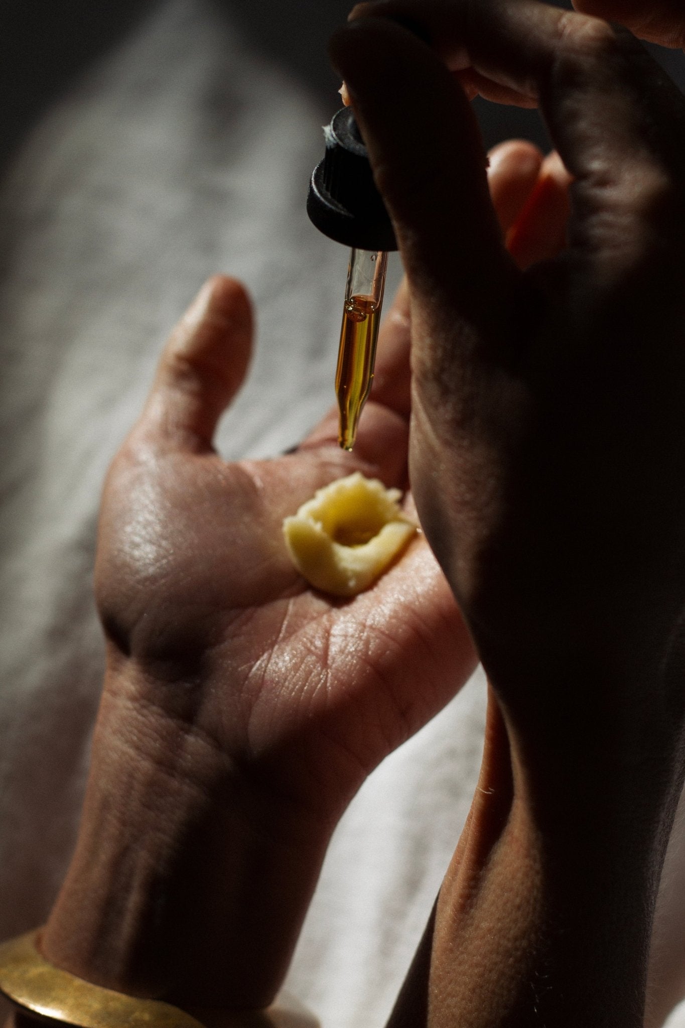 A hand holds a dollop of cream as Emanations Profound Face Oil by Summer Solace Tallow—featuring tallow, emu oil, champa, tuberose & white ginger lily—is dispensed from a dropper above, sunlight casting gentle shadows.