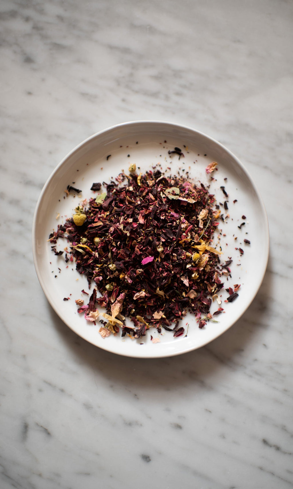 On a marble surface, a white plate displays COAST | Antioxidant Protection Herbal Infusion by PLANTWALK—an antioxidant blend of dried tea leaves and flower petals in vibrant red, purple, yellow, and green.