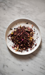 On a marble surface, a white plate displays COAST | Antioxidant Protection Herbal Infusion by PLANTWALK—an antioxidant blend of dried tea leaves and flower petals in vibrant red, purple, yellow, and green.