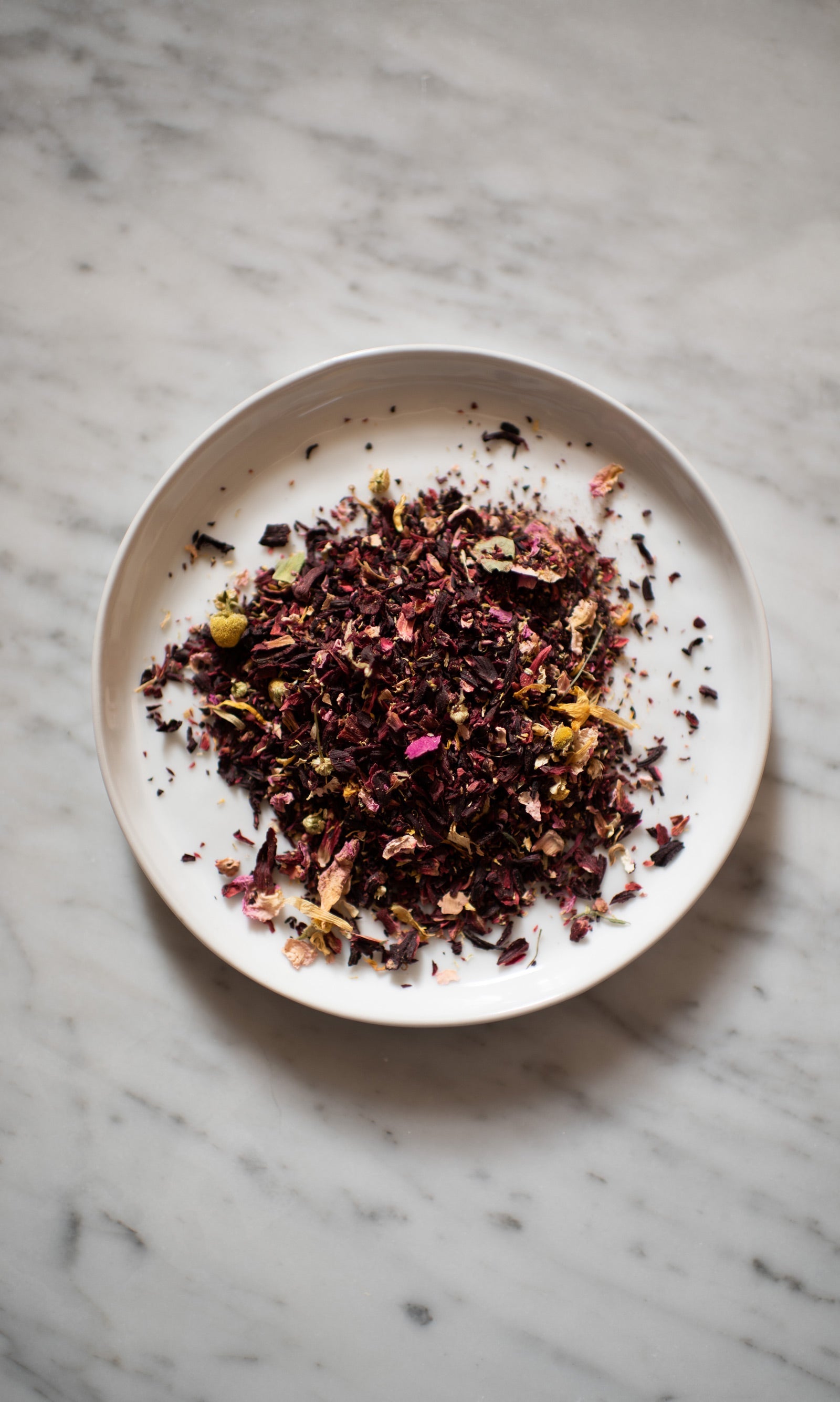 On a marble surface, a white plate displays COAST | Antioxidant Protection Herbal Infusion by PLANTWALK—an antioxidant blend of dried tea leaves and flower petals in vibrant red, purple, yellow, and green.