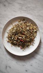A white plate on marble holds a loose pile of PLANTWALK’s FOREST | Healthy Hair & Skin Herbal Infusion—dried herbs and flower petals crafted to support hair and nail strength with natural silica benefits.