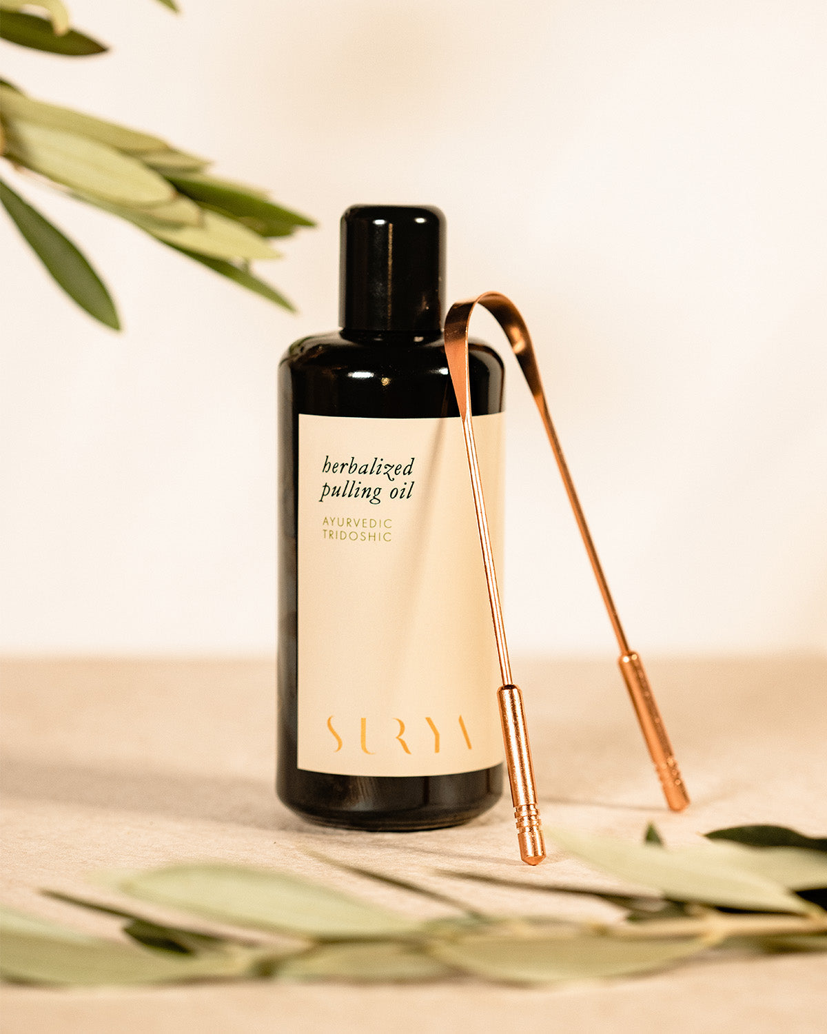 A dark glass bottle of Surya Herbalized Pulling Oil stands upright on a light surface next to a copper tongue scraper, reflecting Ayurveda-inspired dental care, with blurred leaves in the foreground and background.