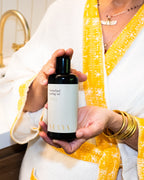 A person in white and yellow robes holds a dark bottle of Surya Herbalized Pulling Oil, showcasing Ayurvedic dental care, with gold bracelets and a visible gold necklace.