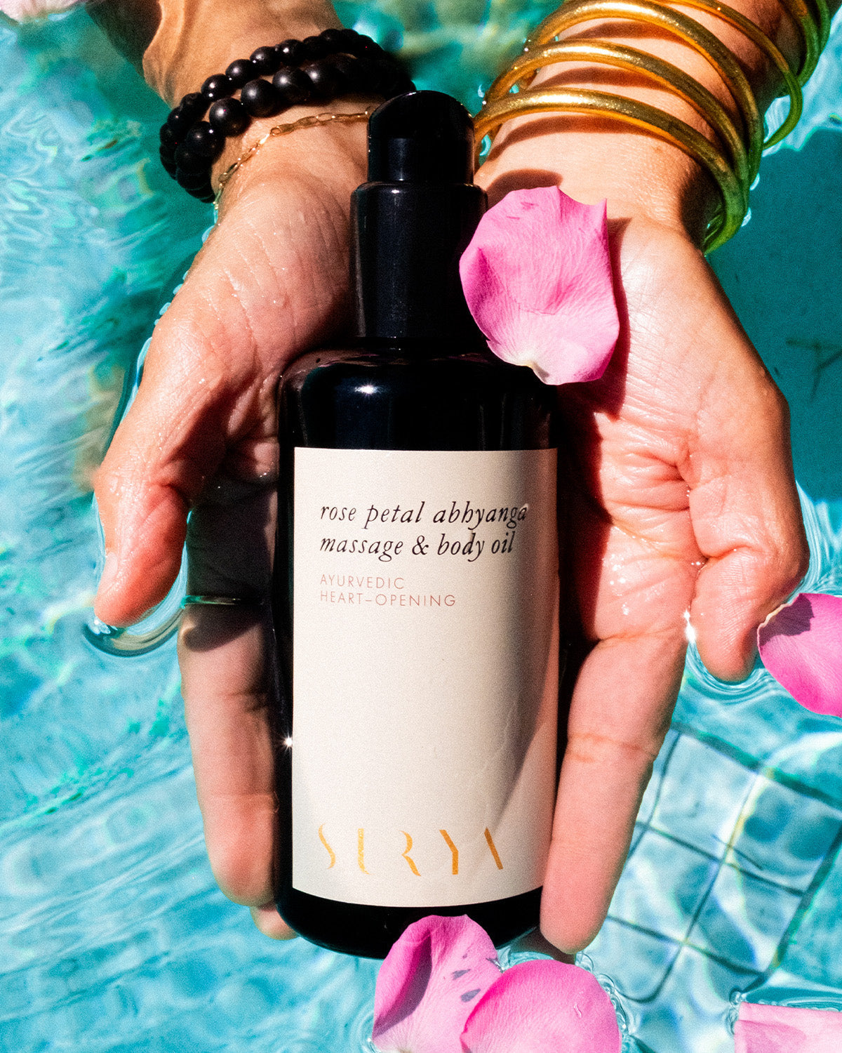 Hands over water hold a dark Surya Rose Petal Body Oil bottle, surrounded by pink rose petals and spa vibes—perfect for a relaxing, rejuvenating massage.
