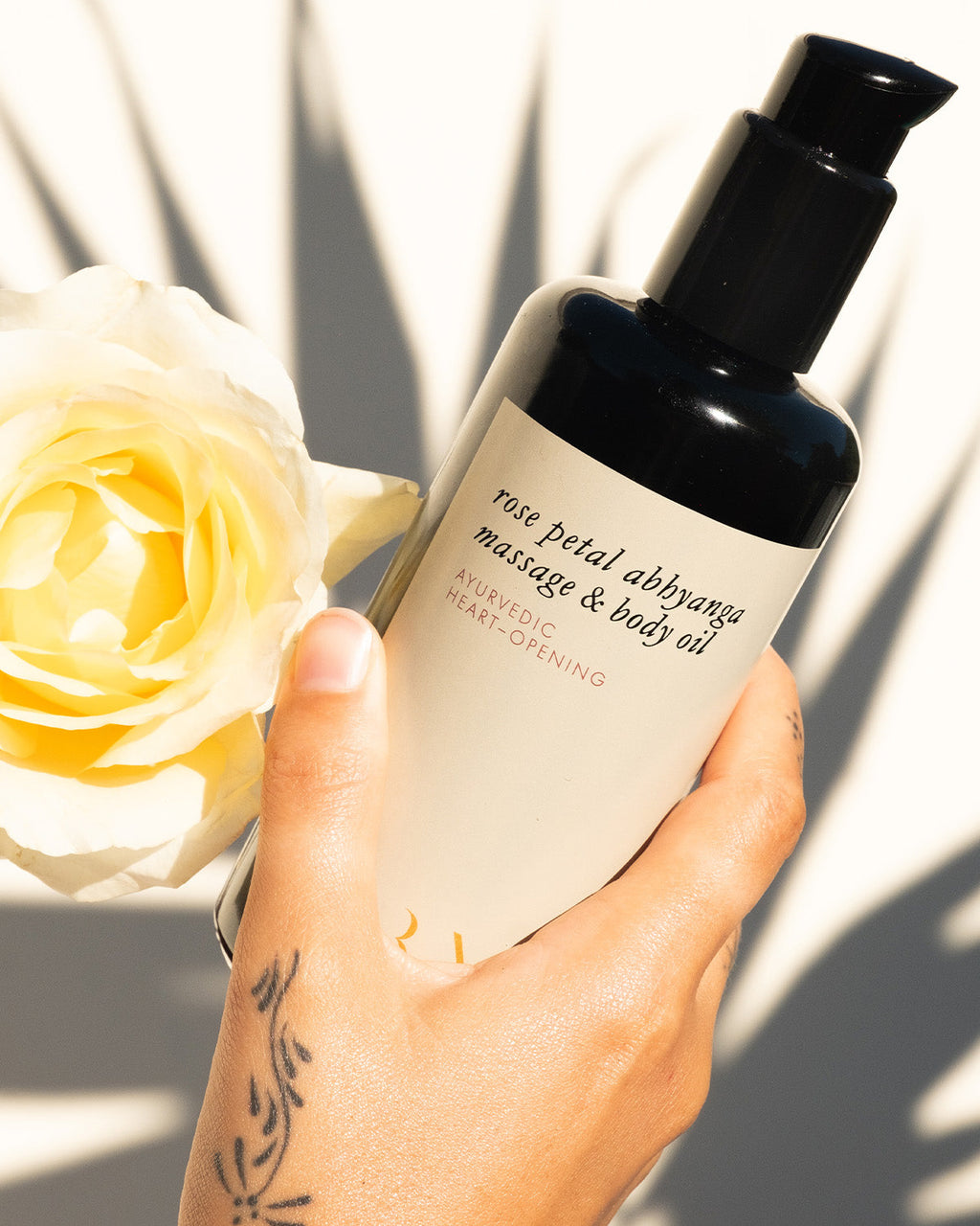 A hand with a floral tattoo holds Surya's Rose Petal Body Oil beside a pale yellow rose, as bright sunlight casts shadows on a white background.