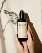 A person holds a dark glass bottle of Surya Slimming Massage Oil—ideal for lymphatic self-massage or cellulite reduction—against a beige background with a plant's shadow.