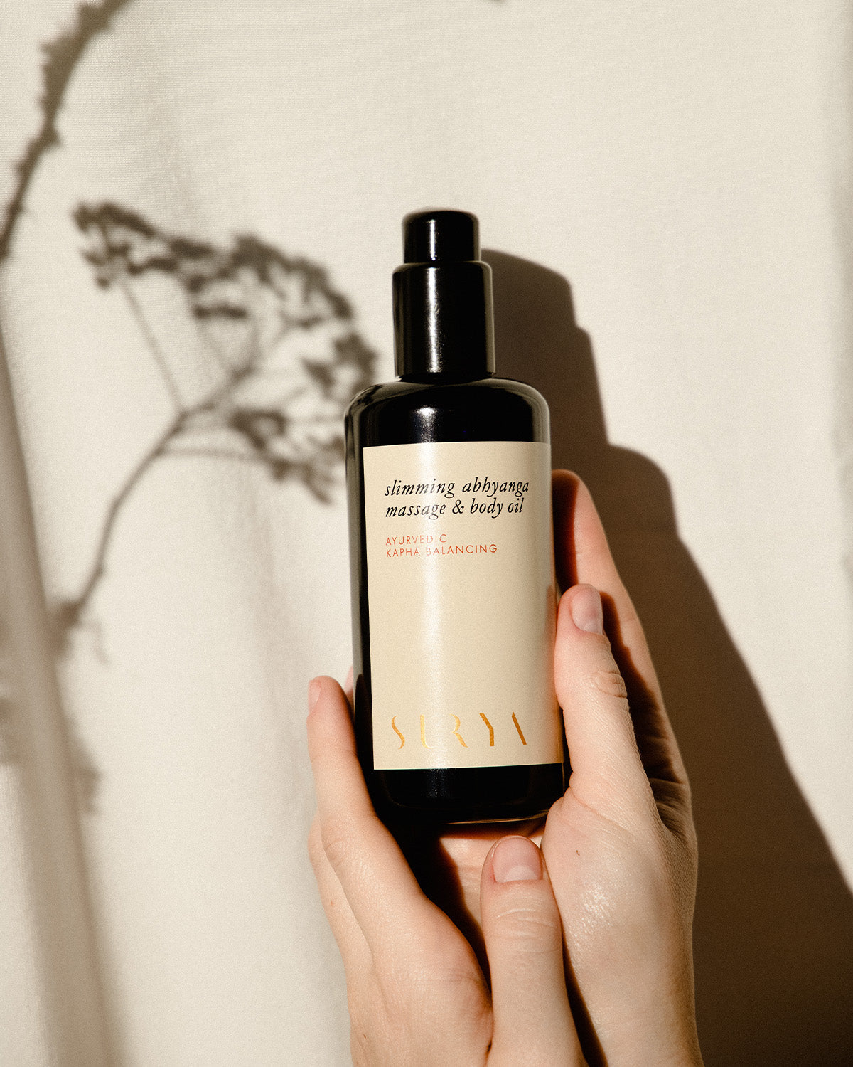 A person holds a dark glass bottle of Surya Slimming Massage Oil—ideal for lymphatic self-massage or cellulite reduction—against a beige background with a plant's shadow.