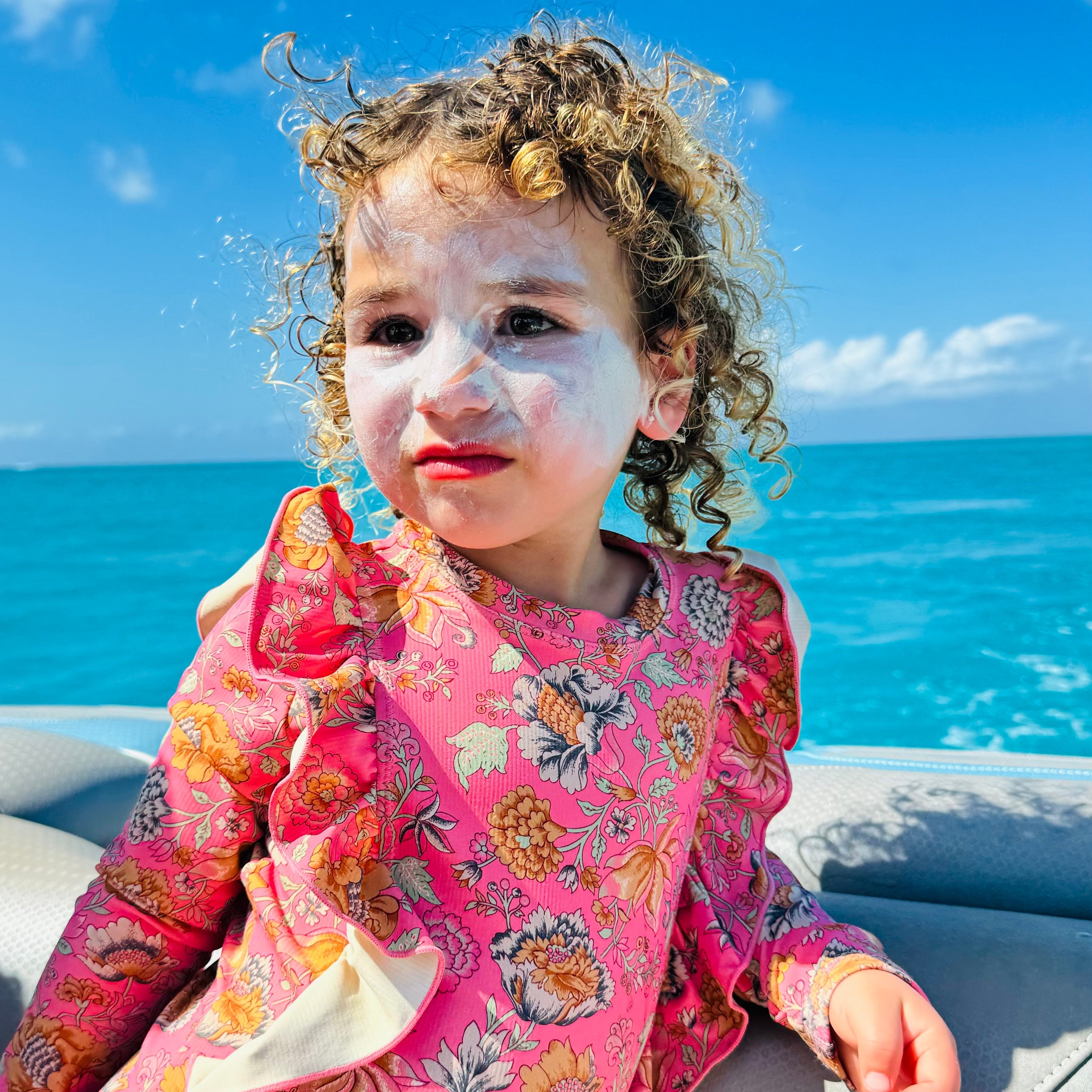 A young child with curly hair and Wild Mamas Sunscreen- Wild Bison Tallow Balm on their face sits on a boat in a pink floral dress, with the ocean and blue sky in the background.