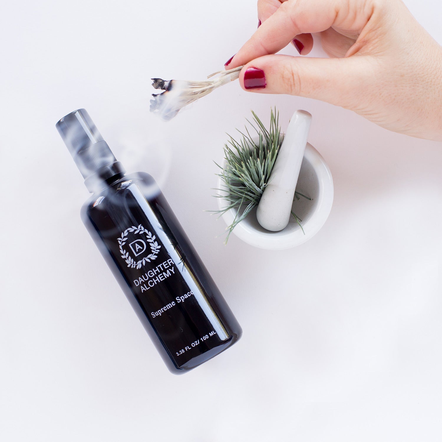 A hand with red nail polish holds smoldering sage above a black "Supreme Space® Botanical Protective Spray" bottle from Daughter Alchemy and a white mortar with a small green air plant, evoking ritual and renewal.