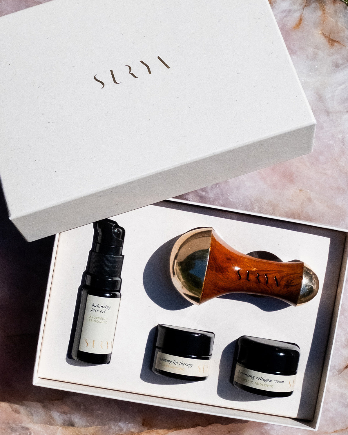 The Surya Skincare Discovery Set features a cream-colored lid labeled "SURYA" and includes a spray bottle, Kansa wand facial massage tool, and two small jars, all neatly arranged for an Ayurvedic skincare experience.