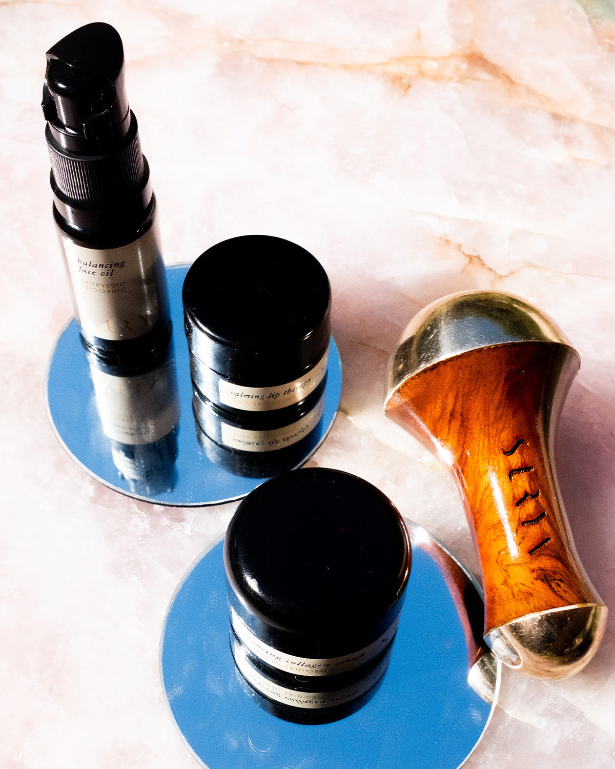 Three Surya Skincare Discovery Set containers and a Kansa wand rest on circular mirrors atop pink marble. Reflected light highlights the Surya set and Ayurvedic massager, creating an elegant arrangement.