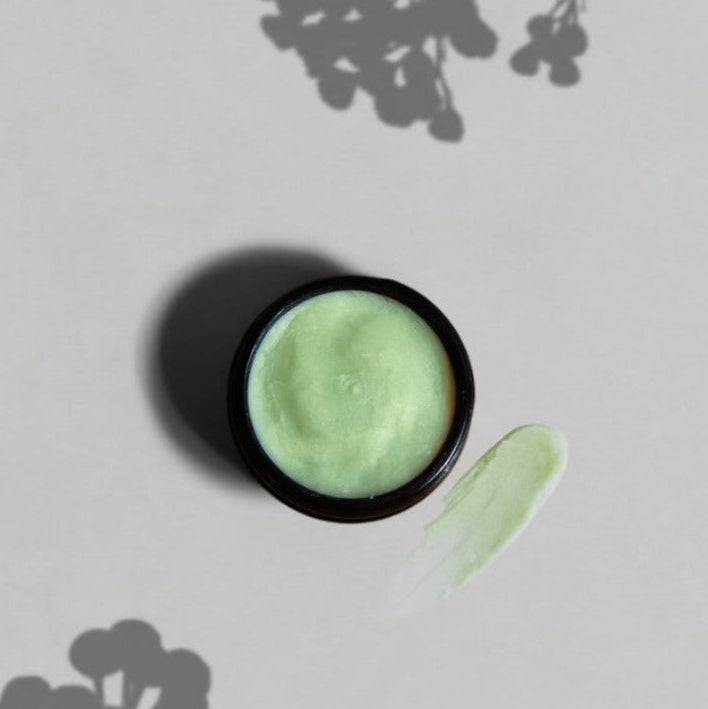 The Wild Mamas Ancestral Blue 2.4oz Wild Bison Tallow Balm, in a small round container with light green cream, rests on a pale surface beside a smear of balm and soft plant shadows in the background.