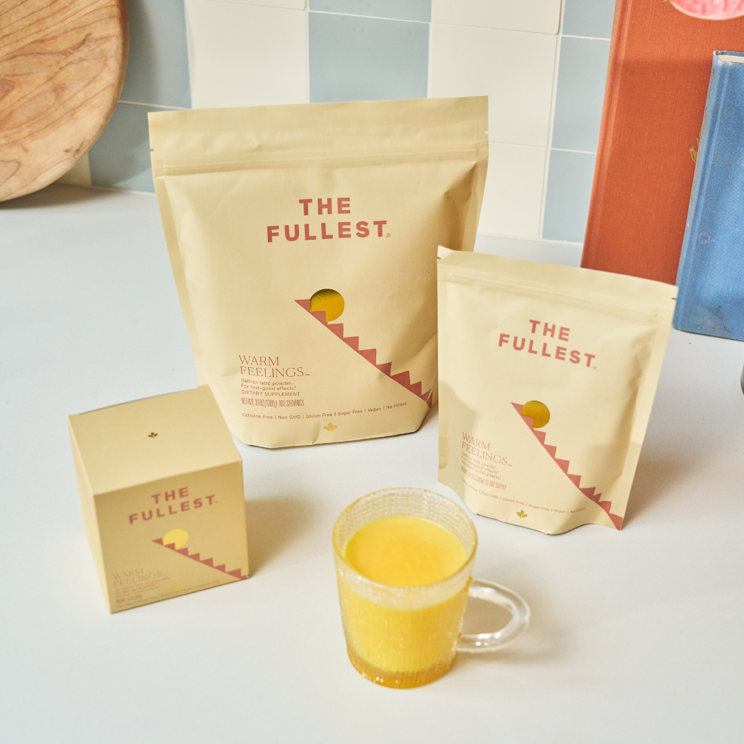 A glass mug of caffeine-free Saffron Latte sits on a white countertop in front of three beige boxes labeled "The Fullest Saffron Latte 10 Single-Serving Sachets," with a tiled backsplash behind them.