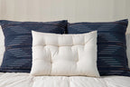 Three pillows are arranged on a bed. Two large, dark blue pillows featuring horizontal stripes sit at the back, with a smaller tufted white pillow positioned in front. The white bedding is accented by a Shepherd's Dream Wool Contour Sleep Pillow for enhanced neck support, set against a light-colored wall.