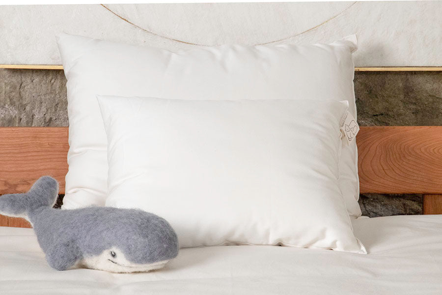 A neatly made bed showcases Shepherd's Dream Child's Sleep Pillow, crafted with natural wool, alongside a small whale-shaped stuffed toy on top. The wooden bed frame pairs harmoniously with the stone-textured wall, fostering a serene and healthy bedroom atmosphere.